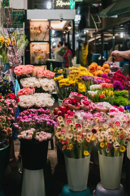 A vibrant flower stall at an outdoor market, showcasing an array of fresh flowers arranged in bouquets and buckets. Prominent among them are pink peonies, white roses, and colorful tulips, all display