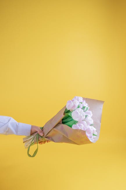 A person extending their hand, dressed in a white shirt, holds a bouquet of fresh pink tulips wrapped in light brown paper with a green ribbon, presenting it against a bright yellow background. The bo