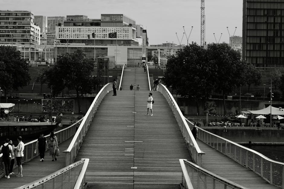 A black-and-white photograph of the Millennium Bridge in London, showing a wide wooden walkway with mice rails on either side, populated by pedestrians walking and standing. The bridge's curved white 