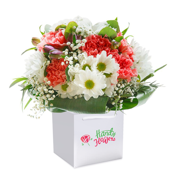 Redolent of artistry and freshness, this vibrant bouquet is presented in an immaculate white gift bag, capturing the refined yet welcoming spirit of De Beauvoir Town. A lush gathering of coral and peach carnations, their ruffled petals dancing in subtle layers of colour, forms the heart of the arrangement, surrounded by dainty white daisies with bright yellow-green centres that glimmer under soft daylight, reminiscent of the early mornings along Kingsland Road. Wispy accents of white baby's breath are thoughtfully scattered, weaving through the more substantial blooms to impart a sense of light and movement. Nestled on the left, a few closed buds tinged with a soft purple-pink shade bring a playful intrigue to the mix, echoing the creative charm of the neighbourhood's art studios. Abundant glossy greenery arcs around the base, offering both visual depth and a polished finish, much like the manicured gardens in De Beauvoir's hidden squares. The arrangement sits upright in a sleek gift bag with sturdy white ribbons, symbolizing both urban convenience and elegant presentation. Photographed against an unbroken white background, the bouquet stands as a testament to local expertise-ideal for next-day delivery throughout De Beauvoir Town and a thoughtful choice for birthdays, anniversaries, or special serendipitous surprises. Every detail embodies the blend of creativity and care present in De Beauvoir Town Florist's approach to contemporary floristry.
