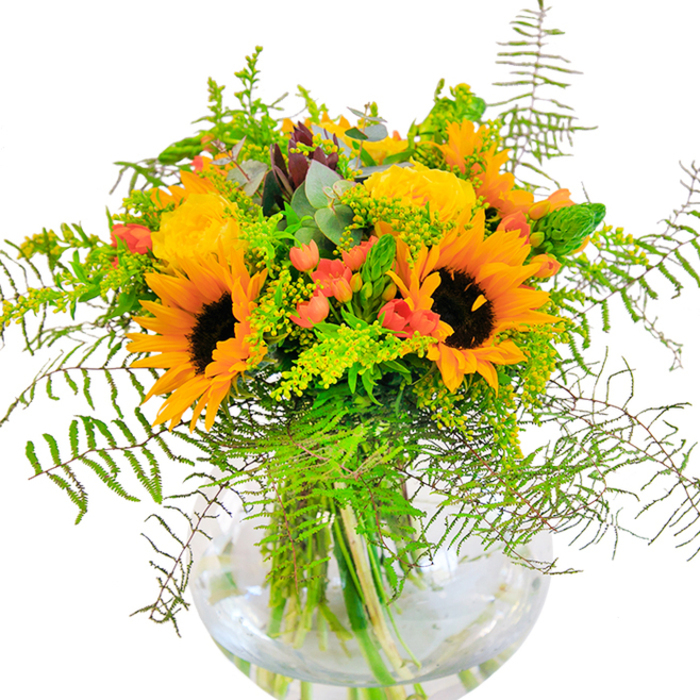 Radiant and uplifting, this bouquet is a lively tribute to the character of De Beauvoir Town: bold, abundant, and seamlessly tied to the warmth of the neighbourhood. Sunflowers dominate the arrangement, their broad faces and golden-orange petals recalling the brightness of a stroll down De Beauvoir Square in midsummer. Interwoven with these cheerful blooms, buttery yellow garden roses present a classic, swirling form-each layer of petals amplifying the bouquet's sense of fullness and joy. Red Hypericum berries dot the composition, their shimmering skins providing both visual and textural intrigue among the greenery. Feathery fern fronds spill outward, mingling with light filler blossoms and slender green accents that add a fresh, wild texture reminiscent of De Beauvoir Town's vibrant urban gardens. The base is a flawless, fishbowl-style glass vase that holds every stem in crisp, clear water, lending the arrangement a modern touch. Flecks of bluish eucalyptus bring a soothing undercurrent, rounding out the scene with a hint of woodland calm. The scent is delicately floral, with hints of earthy green, filling any room with an uplifting ambience. Perfectly considered for birthdays, milestone occasions, or simply as a radiant centerpiece on a De Beauvoir Town dining table, this bouquet channels both the energy and serenity of the local area.