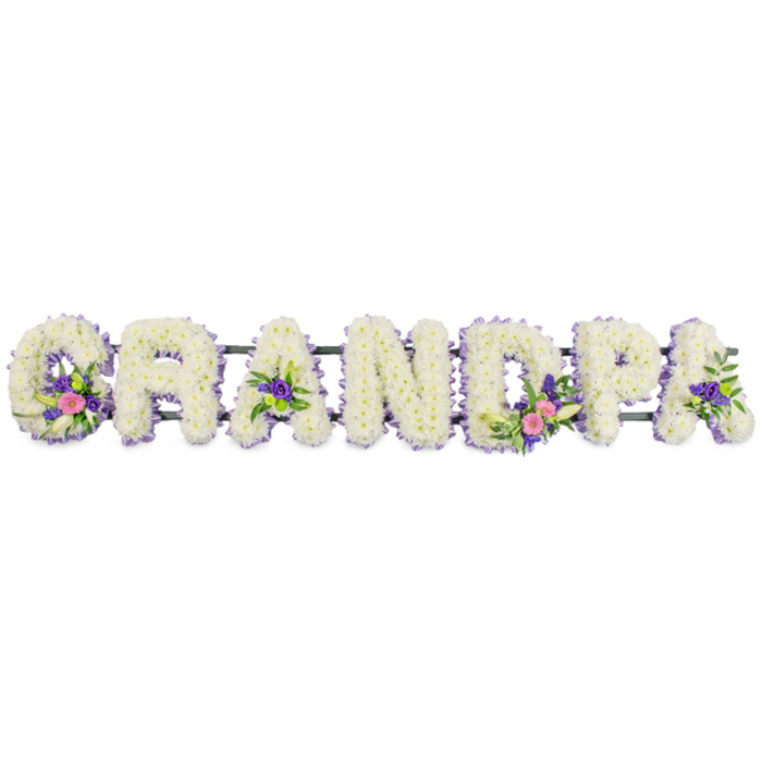 In this carefully lit photograph, the name GRANDPA is recreated in flowers, each letter formed as a substantial three-dimensional design set horizontally across a stark white background. The seven capital letters are built from dense mats of small white chrysanthemums or similar traditional funeral blooms, packed closely together so that their many fine petals merge into a velvety, cushion-like surface. This expanse of pure white suggests peace, reflection, and the gentle respect often associated with saying goodbye to a grandparent in the De Beauvoir Town community. Encircling every letter is a narrow trim of lilac and purple blossoms, likely statice or heather, providing a subtle yet effective outline that defines the shapes and adds a cool hint of colour, reminiscent of quiet evening light along De Beauvoir Square. Within selected letters, the florist has arranged small yet expressive accent sprays: the 'G' carries a cluster of vivid pink and fuchsia blooms intertwined with a slender white lily-like flower and touches of deep purple; the first 'A' holds a more modest but equally tender grouping; the 'D' showcases another prominent spray combining pink, white, and purple; and the second 'A' includes both coloured flowers and some bright green button chrysanthemums or similar foliage. These accents punctuate the tribute with pockets of character, making the piece feel personal rather than purely formal. A slim, dark green frame runs unobtrusively along the back of the letters, supporting them in a neat line ready for placement at a funeral service, wake, or graveside. The overall mood conveyed is one of loving remembrance and quiet gratitude, a floral farewell that honours a grandfather's life and presence within his family and neighbourhood.